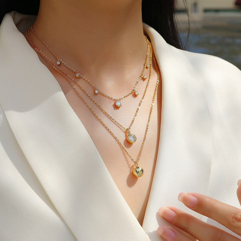 Woman wearing multiple gold necklaces with various pendants against a blurred outdoor background